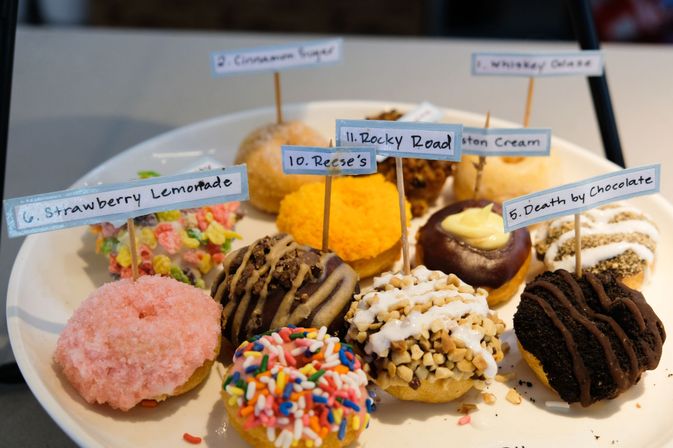 Assorted mini gourmet donuts on a white plate at a bakery counter, colorful toppings and handwritten flavor flags (Strawberry Lemonade, Reese's, Rocky Road, Death by Chocolate), with sprinkles, nuts and chocolate drizzles