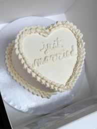 Heart-shaped white buttercream wedding cake with ruffled piping and 'just married' script, set on a silver board inside a bakery box.