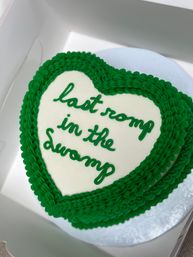 Heart-shaped white frosted cake with thick green piped border and green cursive icing reading "last romp in the swamp" tucked inside a white pastry box