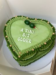 Playful heart-shaped green birthday cake with piped buttercream ruffles, gold dragees, an olive skewer topper and white lettering reading A TINI BIT OLDER