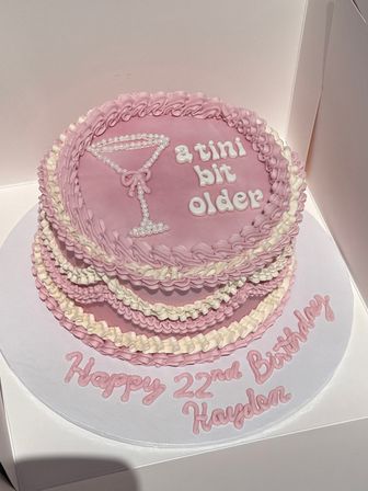 Pink two-tier birthday cake with piped ruffle borders, white pearl‑dot martini glass decoration and icing that reads “a tini bit older,” sitting on a white cake board with a handwritten birthday message.