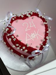 Heart-shaped pink fondant celebration cake with white "24 A*K" lettering, red ruffled buttercream border, baby's-breath flowers and white satin ribbon bows on a silver cake board.