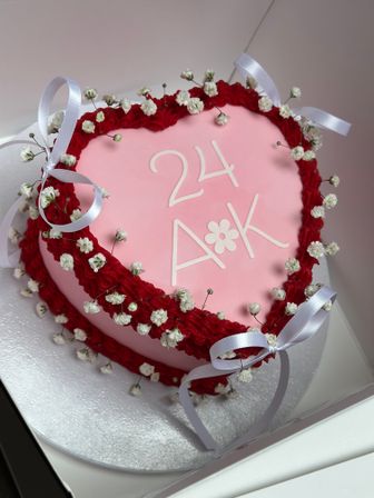 Heart-shaped pink fondant celebration cake with white "24 A*K" lettering, red ruffled buttercream border, baby's-breath flowers and white satin ribbon bows on a silver cake board.