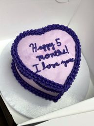 Heart-shaped light purple celebration cake with deep purple ruffled frosting and piped message “Happy 5 months! I love you” on a silver cake board inside a white box