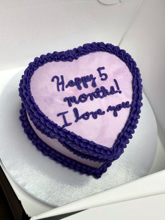 Heart-shaped light purple celebration cake with deep purple ruffled frosting and piped message “Happy 5 months! I love you” on a silver cake board inside a white box