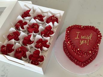Heart-shaped red engagement cake reading "I said yes!" next to a box of matching red buttercream cupcakes with gold pearls on a marble countertop