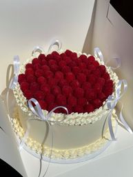 Round white buttercream celebration cake topped with a dense layer of fresh raspberries, piped cream borders and white satin ribbon bows, photographed inside a pastry box.