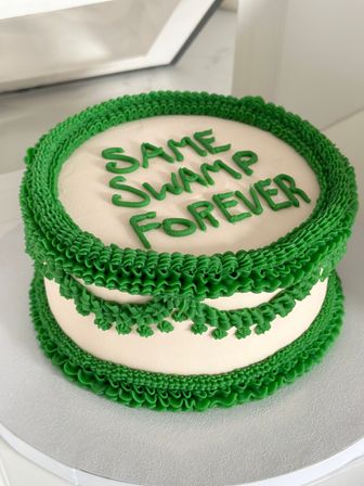 White round cake decorated with vivid green buttercream ruffles and garlands, top piped with the playful message 'SAME SWAMP FOREVER'.