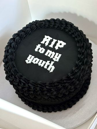 Round black-frosted cake with ruffled piped border and white Gothic lettering reading "RIP to my youth" resting in a white cake box