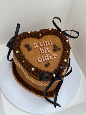 Heart-shaped coffee-themed birthday cake with caramel-brown buttercream, dark chocolate piped border, edible coffee beans and gold dragees, pink lettering reading 'a tini bit older' and black ribbon bows on a white cake board.