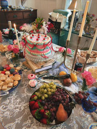Festive party spread on a lace-covered dining table in a cozy dining room: pastel pink-and-teal decorated layer cake on a glass stand, assorted macarons and pastries, cupcakes, a fruit bowl of grapes, strawberries and pear, wine, floral arrangements and lit candelabra.