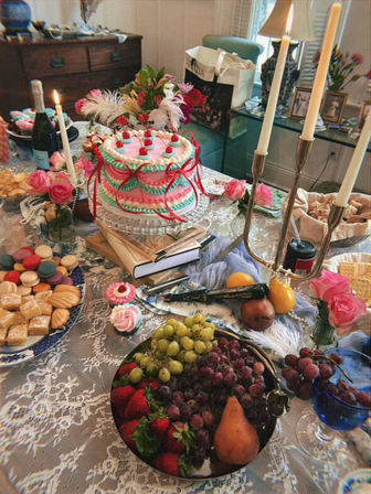 Festive party spread on a lace-covered dining table in a cozy dining room: pastel pink-and-teal decorated layer cake on a glass stand, assorted macarons and pastries, cupcakes, a fruit bowl of grapes, strawberries and pear, wine, floral arrangements and lit candelabra.