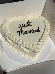 Heart-shaped two-tier white buttercream wedding cake with piped ruffle borders and black "Just Married" script, resting inside a white bakery box