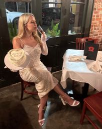 Glam woman in a white beaded dress and long gloves sips champagne while holding a heart-shaped cake that reads 'Almost There' at a cozy brick‑walled restaurant table.