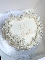 Heart-shaped white buttercream cake with piped ruffles, baby's-breath flowers and pearl sugar decorations, scripted 'til death'—romantic wedding or anniversary dessert.
