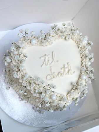 Heart-shaped white buttercream cake with piped ruffles, baby's-breath flowers and pearl sugar decorations, scripted 'til death'—romantic wedding or anniversary dessert.