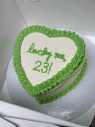 Heart-shaped white cake with bright green piped frosting border and pearl trim, "lucky me, 23!" written in green icing — festive birthday dessert