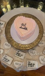 Heart-shaped pastel pink frosted cake on an ornate gold stand reading "til death" in black script, displayed on a wedding dessert table with individually wrapped themed cookies (dates 10.27.24, "Til death do us part", initials V&J) and glass pillar candles.