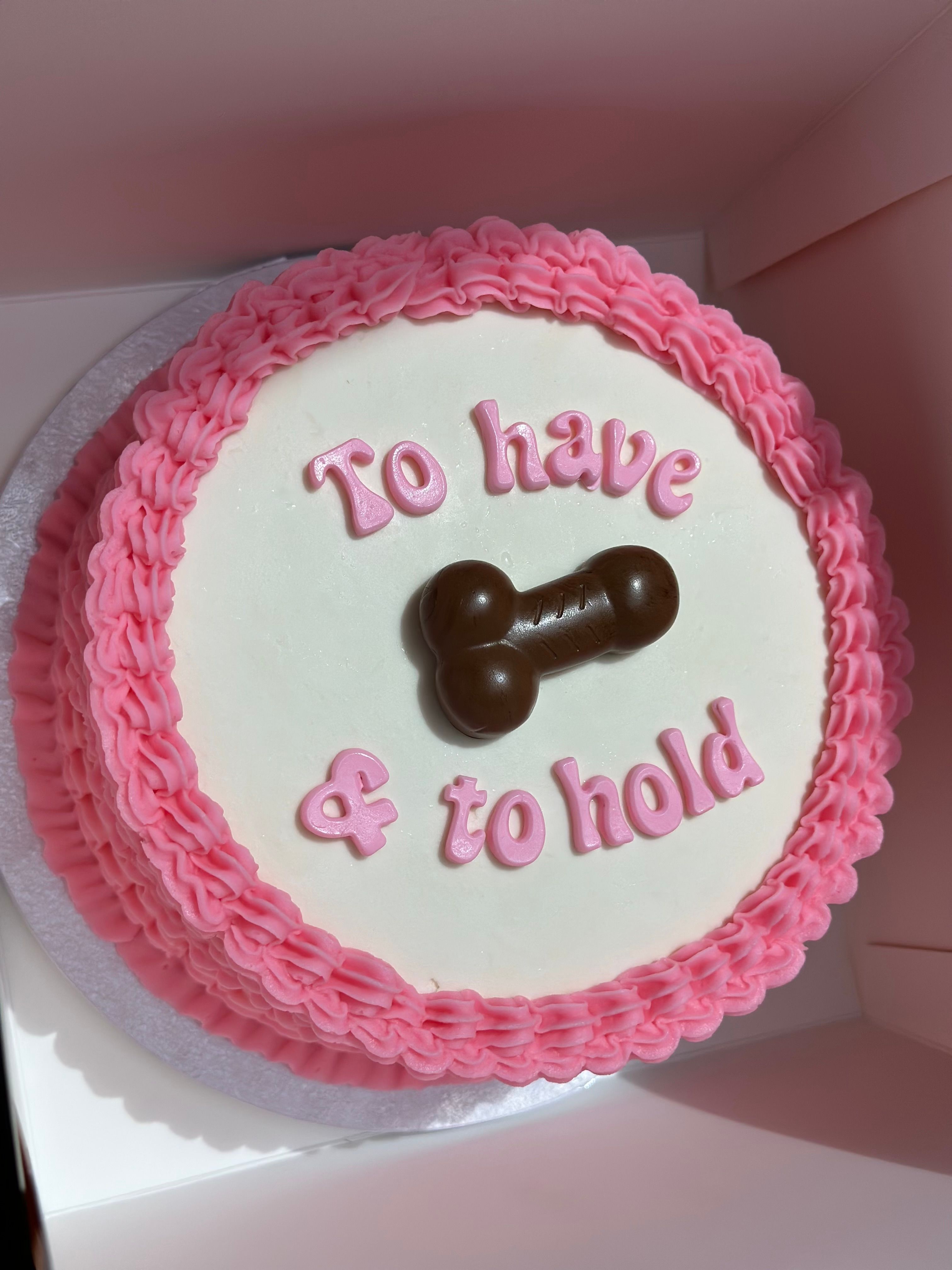 Round white cake with pink ruffled buttercream border, pink letters reading 'To have & to hold' and a chocolate bone-shaped topper — playful wedding/bridal shower cake.