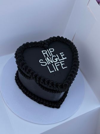 Black heart-shaped cake with piped black border and white icing text reading "RIP SINGLE LIFE," on a white cake board inside a box.