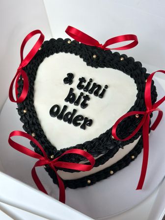 Heart-shaped white frosted cake with black piped icing border, red satin ribbon bows and gold dragees, playful black lettering reading '3 tini bit older'.