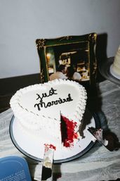 Heart-shaped white wedding cake on a reception table reading "Just Married," with a red velvet slice cut out, cake server with frosting nearby and a framed photo in the background.