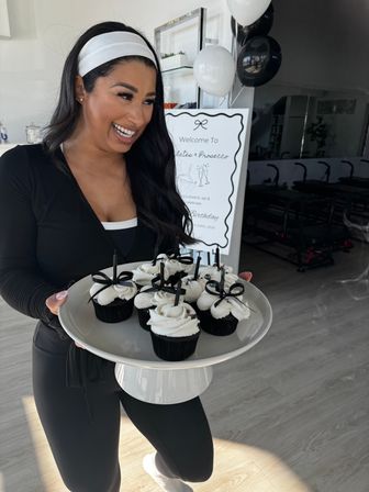 Smiling woman in black workout clothes holding a platter of black cupcakes with white frosting and black ribbon picks inside a bright Pilates studio with balloons and a welcome sign for a Pilates & Prosecco celebration.