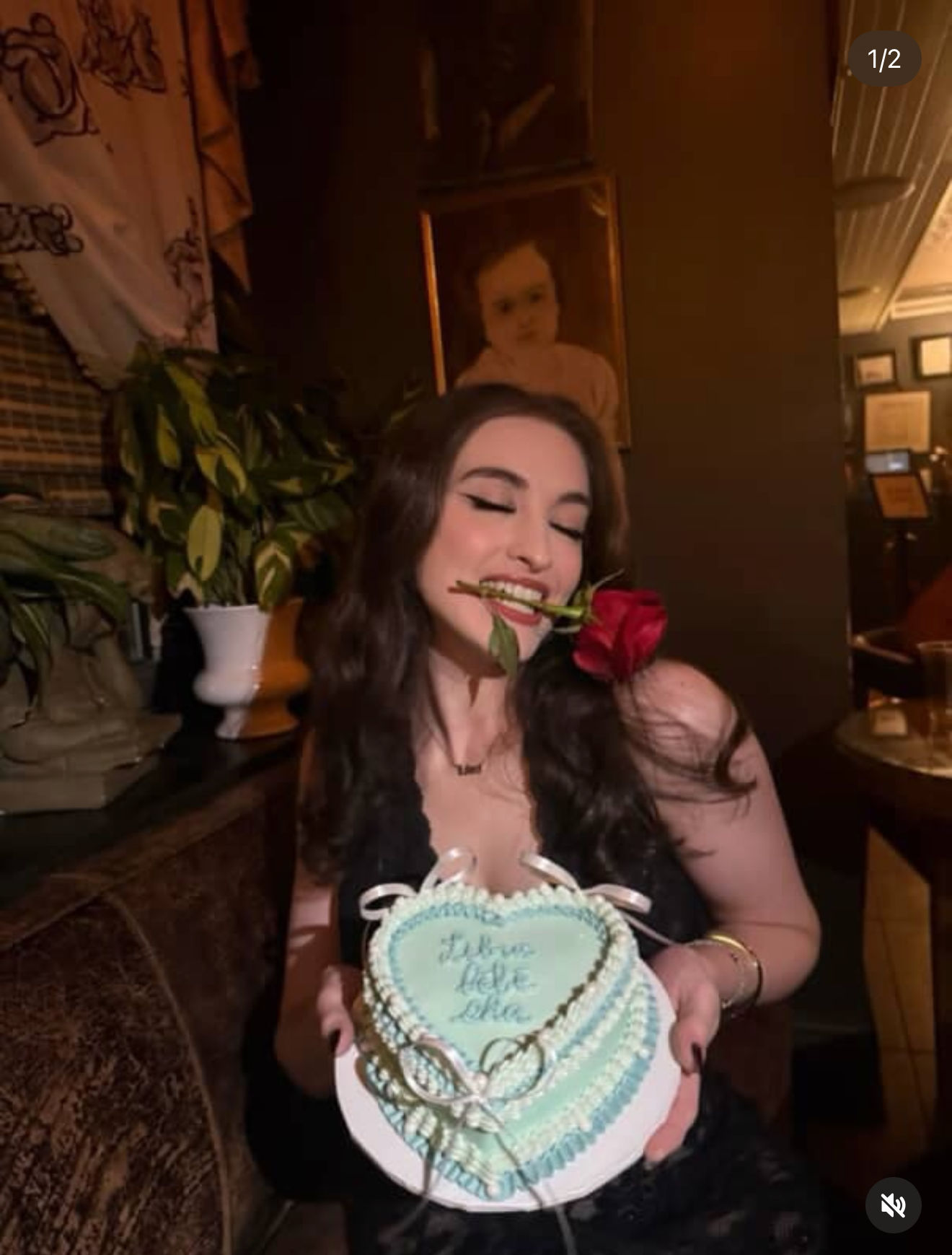 Playful woman in a dimly lit lounge holding a heart-shaped pale blue cake with ribbon accents and cursive icing, a red rose between her teeth, festive celebration vibe.
