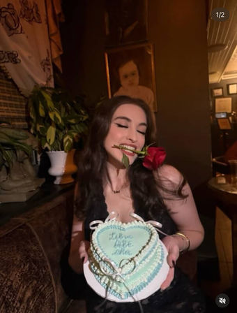 Playful woman in a dimly lit lounge holding a heart-shaped pale blue cake with ribbon accents and cursive icing, a red rose between her teeth, festive celebration vibe.