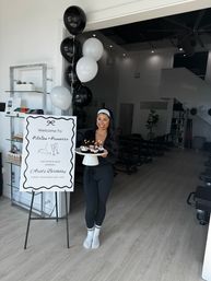 Smiling woman in black activewear holding a cake stand of lit cupcakes next to black-and-white balloons and a welcome sign for a Pilates birthday event inside a bright pilates studio with reformer machines.