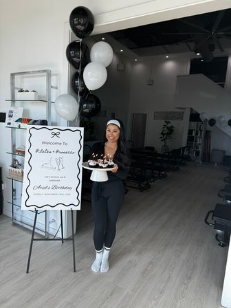 Smiling woman in black activewear holding a cake stand of lit cupcakes next to black-and-white balloons and a welcome sign for a Pilates birthday event inside a bright pilates studio with reformer machines.