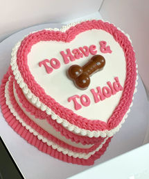 Heart-shaped pink and white frosted cake with ruffled borders, a chocolate penis topper and playful pink lettering reading 'To Have & To Hold', novelty bachelorette/bridal shower cake.