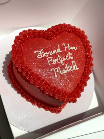 Heart-shaped red cake with ruffled icing border, sparkly red frosting and white piped message "Found Her Perfect Match" inside a white cake box