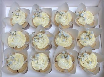 Box of 12 vanilla cupcakes with white buttercream swirls, pearl sprinkles and tiny baby's-breath flowers in tulip parchment liners — elegant wedding-style desserts.