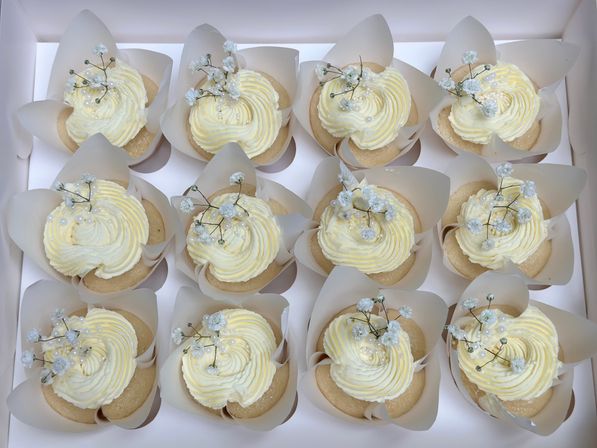 Box of 12 vanilla cupcakes with white buttercream swirls, pearl sprinkles and tiny baby's-breath flowers in tulip parchment liners — elegant wedding-style desserts.