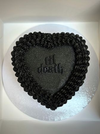 Black heart-shaped cake with sparkly black frosting, piped ruffle border and gothic “til death” lettering, presented on a silver cake board inside a white box.