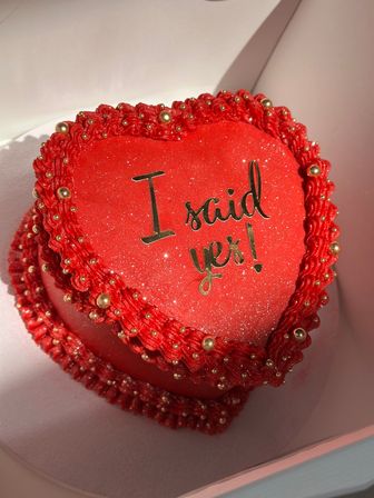 Sparkling red heart-shaped engagement cake with gold bead trim and gold script reading 'I said yes!' in a white box