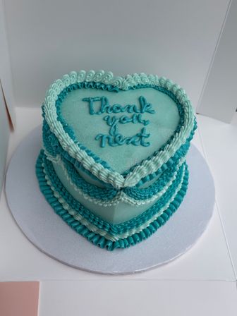 Heart-shaped teal cake with layered buttercream ruffles and piped "thank you next" message, sparkly finish on a white cake board