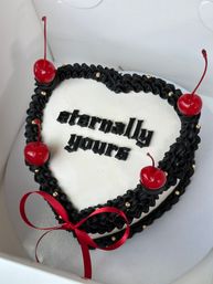 Heart-shaped white cake with black ruffled icing border, gothic black "eternally yours" lettering, glossy red cherries, gold dragees and a red ribbon bow — romantic boxed dessert.