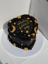 Black two-tier heart-shaped cake with piped black buttercream, decorated with edible gold stars, crescent moons and metallic pearls, gold script reading "witches don't age," on a white cake board