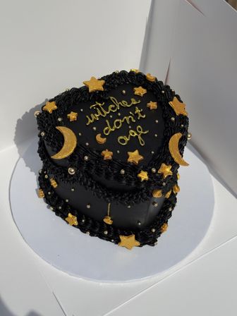 Black two-tier heart-shaped cake with piped black buttercream, decorated with edible gold stars, crescent moons and metallic pearls, gold script reading "witches don't age," on a white cake board