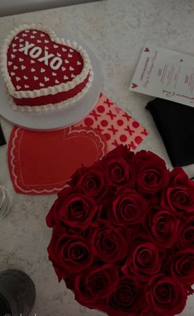 Valentine's Day spread on a light countertop: heart-shaped red cake iced with 'XOXO', heart-patterned napkins and placemats, and a bouquet of deep red roses.