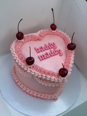 Heart-shaped pink buttercream cake with piped ruffles, shimmery fondant top, pink scripted lettering reading 'baddy maddy' and cherry toppers inside a white cake box