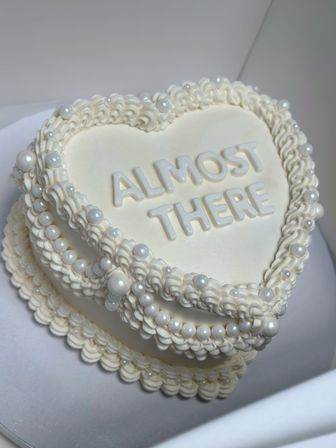 Close-up of heart-shaped white buttercream milestone cake with piped ruffle borders, pearl bead decorations, and raised letters reading 'ALMOST THERE'