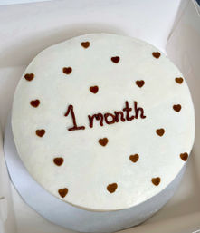 Round white-frosted milestone cake dotted with small chocolate hearts and "1 month" piped in chocolate, on a silver board inside a white cake box.