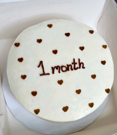 Round white-frosted milestone cake dotted with small chocolate hearts and "1 month" piped in chocolate, on a silver board inside a white cake box.