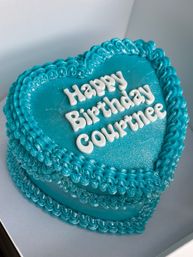 Close-up of a heart-shaped teal birthday cake with shimmering buttercream, decorative piped borders and white lettering spelling a birthday message.