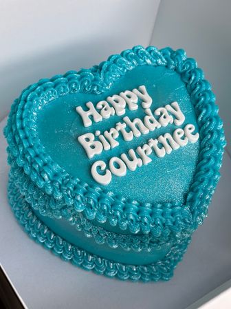 Close-up of a heart-shaped teal birthday cake with shimmering buttercream, decorative piped borders and white lettering spelling a birthday message.
