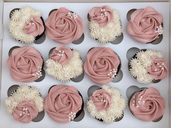 Top-down view of a bakery box of 12 chocolate cupcakes decorated with dusty-pink rose buttercream swirls, white star-piped buttercream accents, and edible pearl sprinkles.
