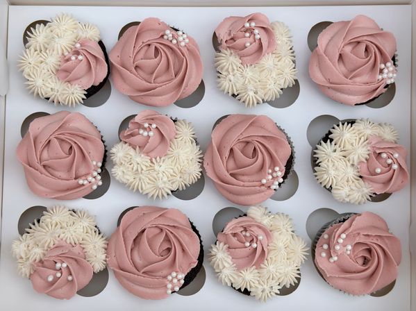 Top-down view of a bakery box of 12 chocolate cupcakes decorated with dusty-pink rose buttercream swirls, white star-piped buttercream accents, and edible pearl sprinkles.